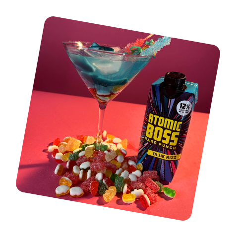 Tilted image with Atomic Boss Blue Rizz and martini glass filled with product with candy and garnish in brightly colored setting