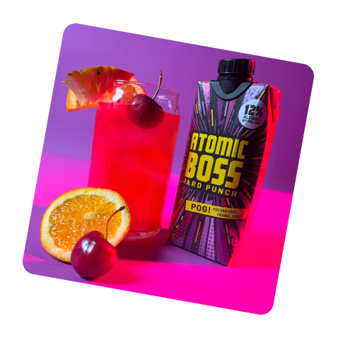Tilted image of Atomic Boss POG! with tall glass filled with product and ice with cherry, orange, and other bits of fruit in still life arrangement in a brightly colored setting